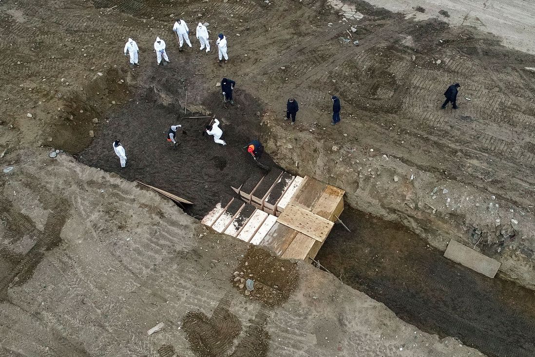 Workers wearing personal protective equipment bury bodies in a trench on Hart Island, in the Bronx. They are standing in the trench, with boxes containing bodies.